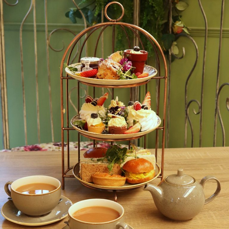 Three-tiered cake stand with assorted finger foods and teacups on a wooden table.