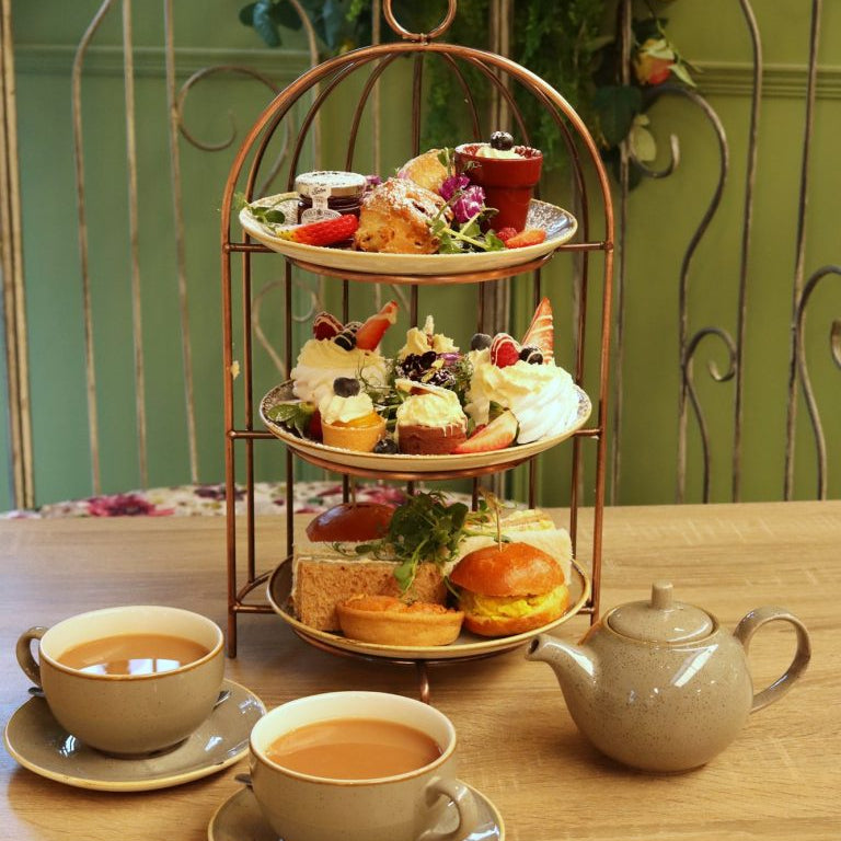 Three-tiered tea stand with assorted finger foods and teacups on a wooden table.
