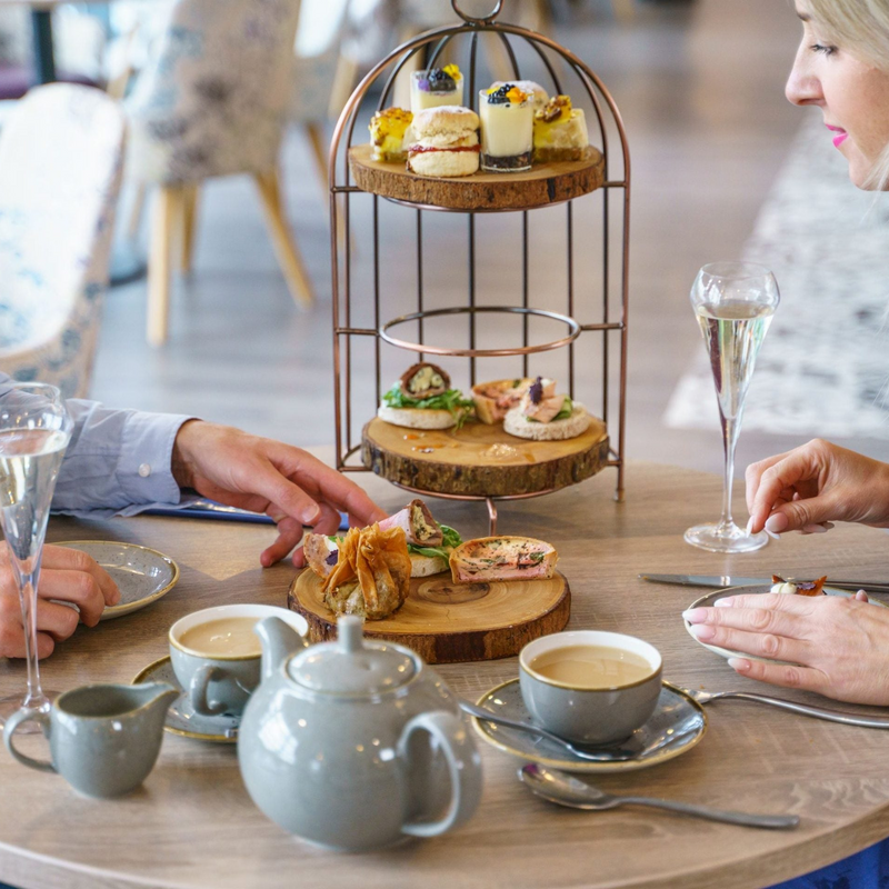 People enjoying afternoon tea with a three-tiered cake stand, teacups, and pastries on a wooden table.